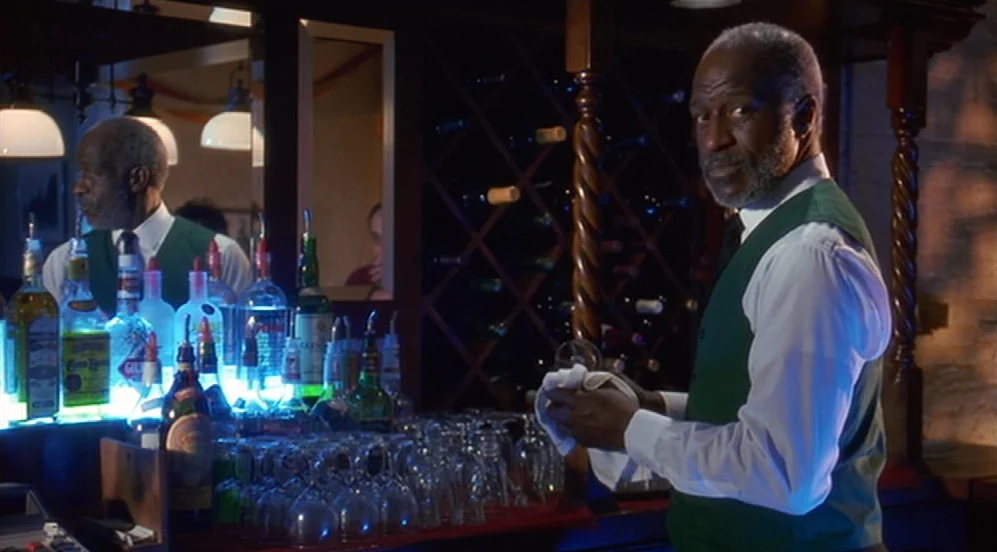 Groundhog Day (1993) movie still: medium — A dimly lit bar counter features a reflective mirror and a rack of wine. An elderly man in a formal vest…