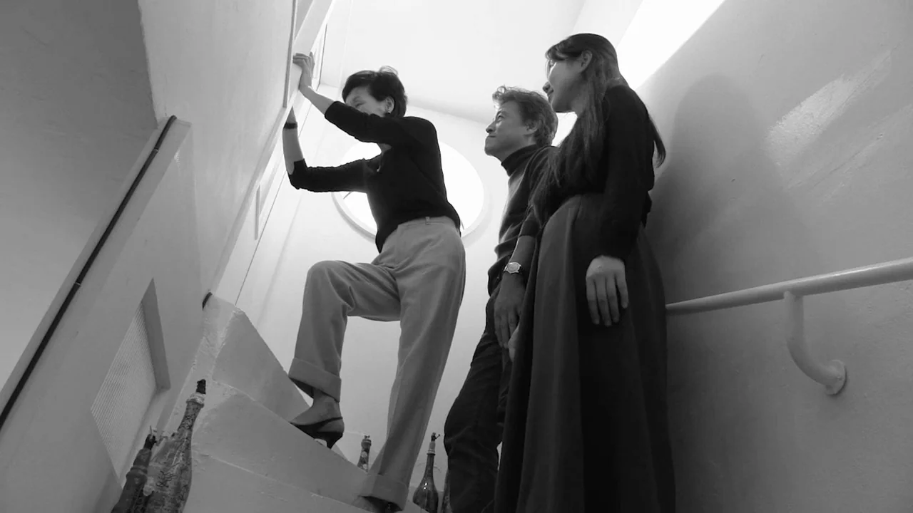 Walk Up (2022) movie still: fullbody — Three people stand on a white staircase beneath a bright circular skylight window. One woman stretches her…