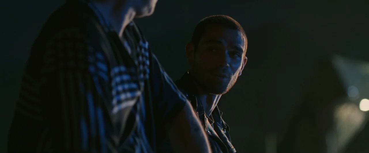 One Fast Move (2024) movie still: medium — A dark outdoor space at night with blue light illuminating the side of a face. Two men are positioned…