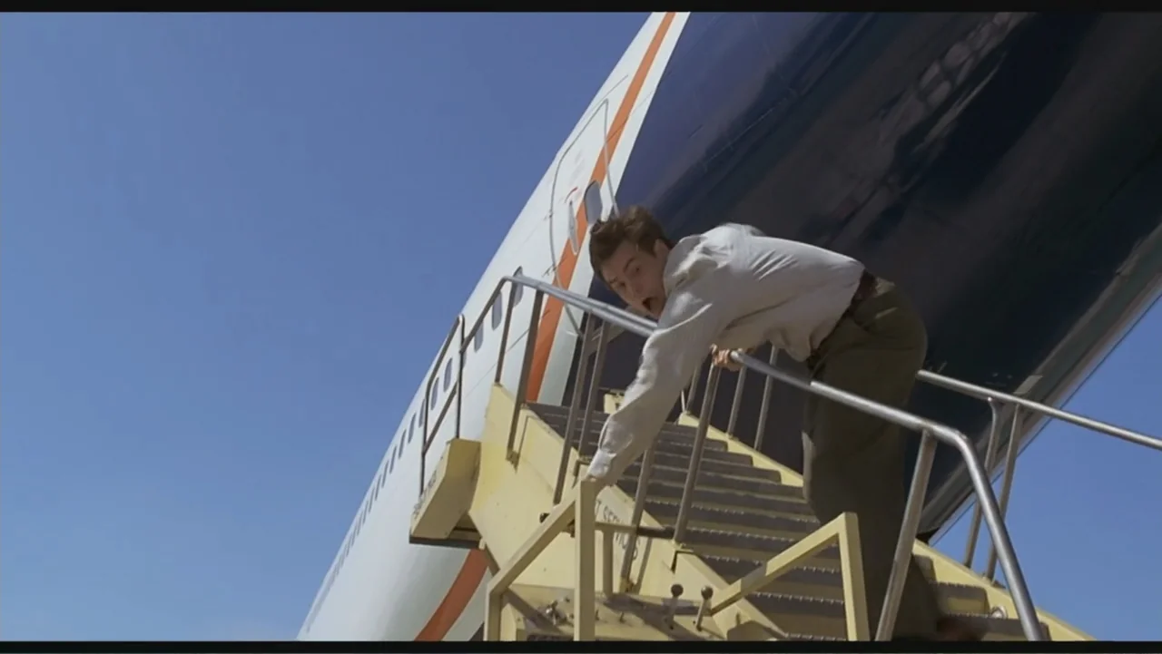 Liar Liar (1997) movie still: medium — An airliner sits on the tarmac under a bright blue cloudless sky. A man in a dress shirt moves down the metal…