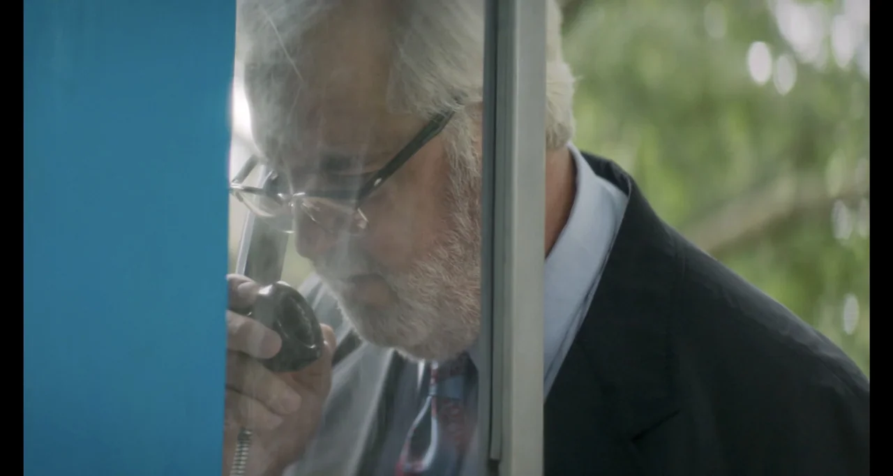 The Poison Rose (2019) movie still: closeup — A glass and blue metal phone booth sits outdoors against a blurry green background. An older man in a…