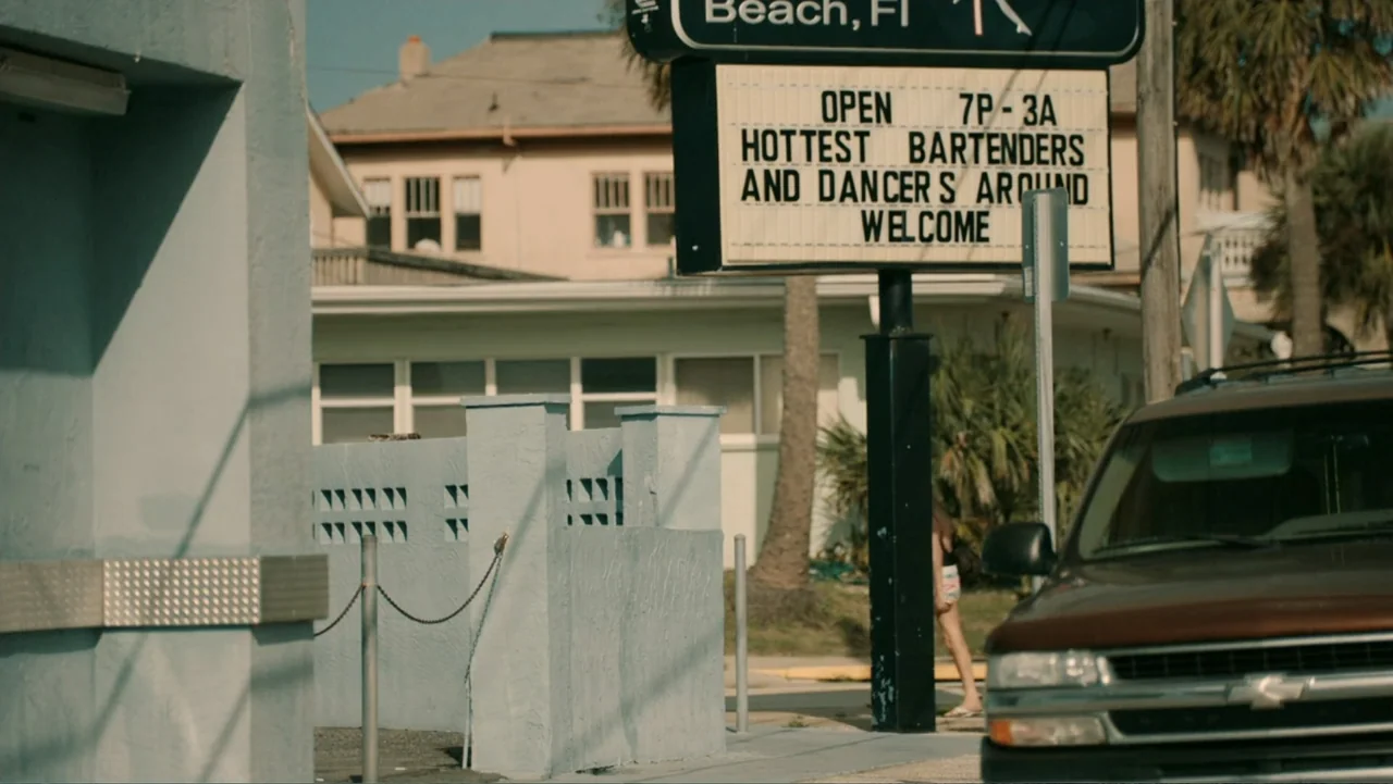 Aileen Wuornos : Mind of a Monster (2020) movie still: wide — A suburban street corner features a marquee sign advertising a bar under clear skies.…