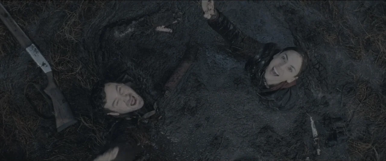 Quicksand (2023) movie still: wide — Two individuals are partially submerged in a deep pit of thick black mud. A rifle lies nearby on the surface as…