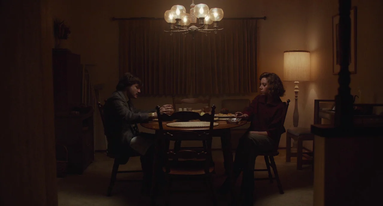 An Evening With Beverly Luff Linn (2018) movie still: wide — A dimly lit dining room features wood furniture, patterned curtains, and a glowing…