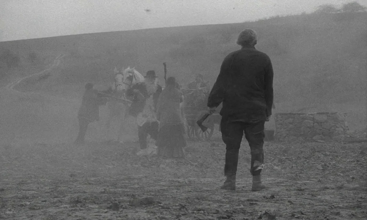The Turin Horse (2011) movie still: wide — A desolate, dusty field on an overcast day under a heavy, featureless sky. A group of people with a…