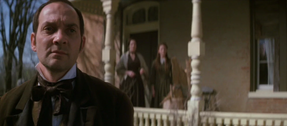Ride With The Devil movie still: medium — A wooden porch of a 19th-century farmhouse features painted balusters and decorative columns. A man stands…