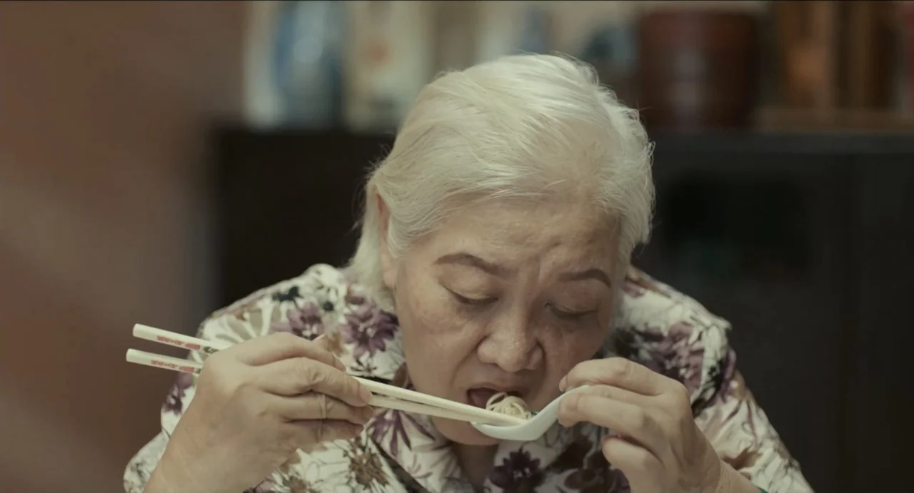 Ramen Shop (2018) movie still: medium — An interior dining area with a blurred dark wooden cabinet in the background. An elderly woman in a floral…