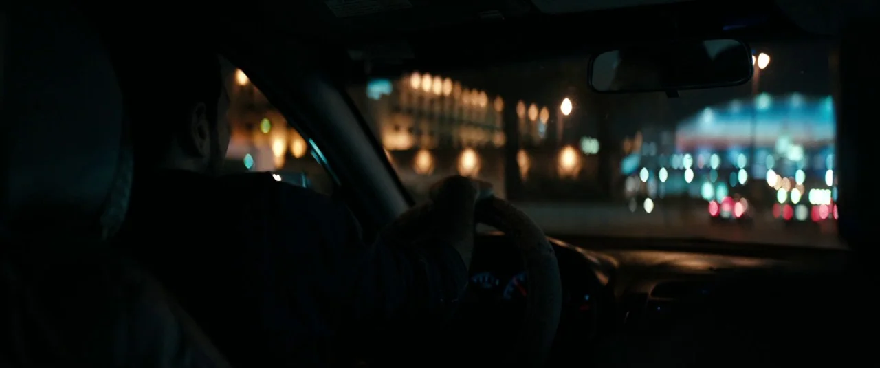 A Hologram for the King movie still: medium — The interior of a moving car at night is cast in deep shadow. A driver is seen in silhouette, steering…
