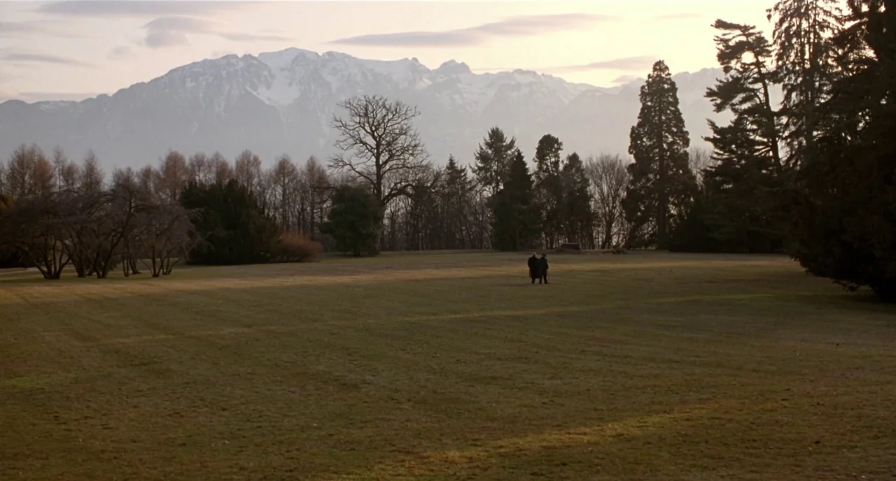 Chaplin (1992) movie still: wide — An expansive grassy meadow sits before a range of tall, snow-capped mountains. Two figures in dark coats stand…