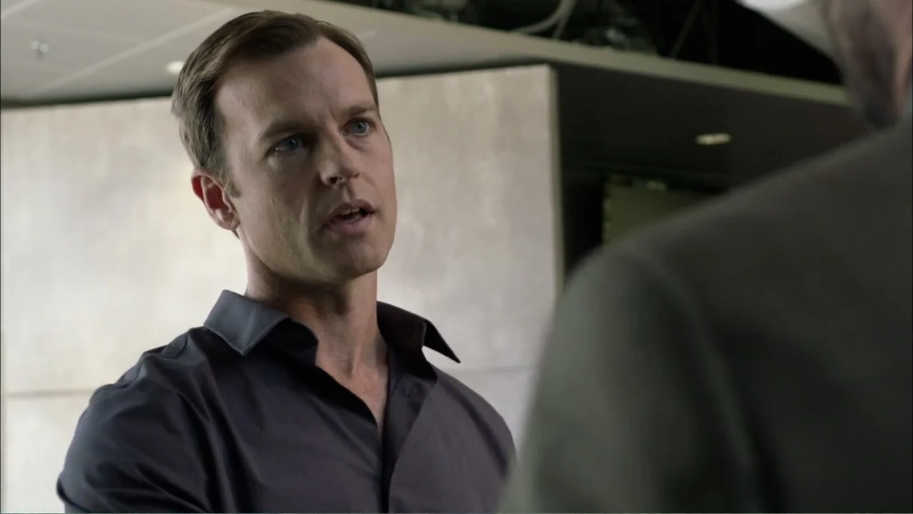 Dark Skies (2013) movie still: medium — An office interior features smooth light-grey walls and a drop ceiling. A man in a dark grey shirt faces his…