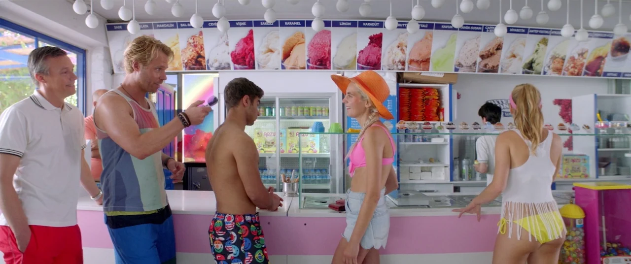 Holiday movie still: wide — An ice cream parlor features a long counter with a menu of colorful flavors displayed. Five people stand in a line…