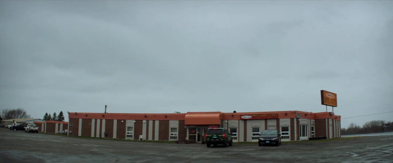 The Silencing (2020) movie still: establishing — A single-story motel building features brown and white brick siding with an orange roof. A dirt…