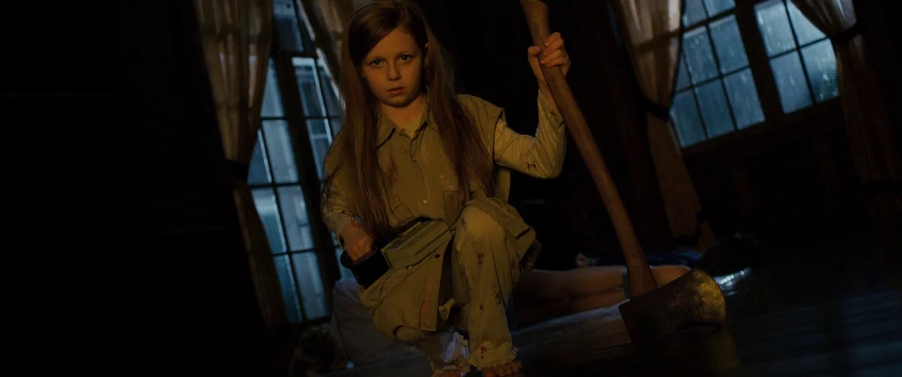 Sinister (2012) movie still: medium — A dark house interior with large windows and tall curtains in the background. A young girl in pajamas crouches…