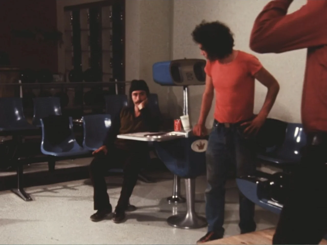 Don't Expect Too Much (2011) movie still: medium — A retro bowling alley seating area features blue plastic molded chairs and a table. A man sits at…