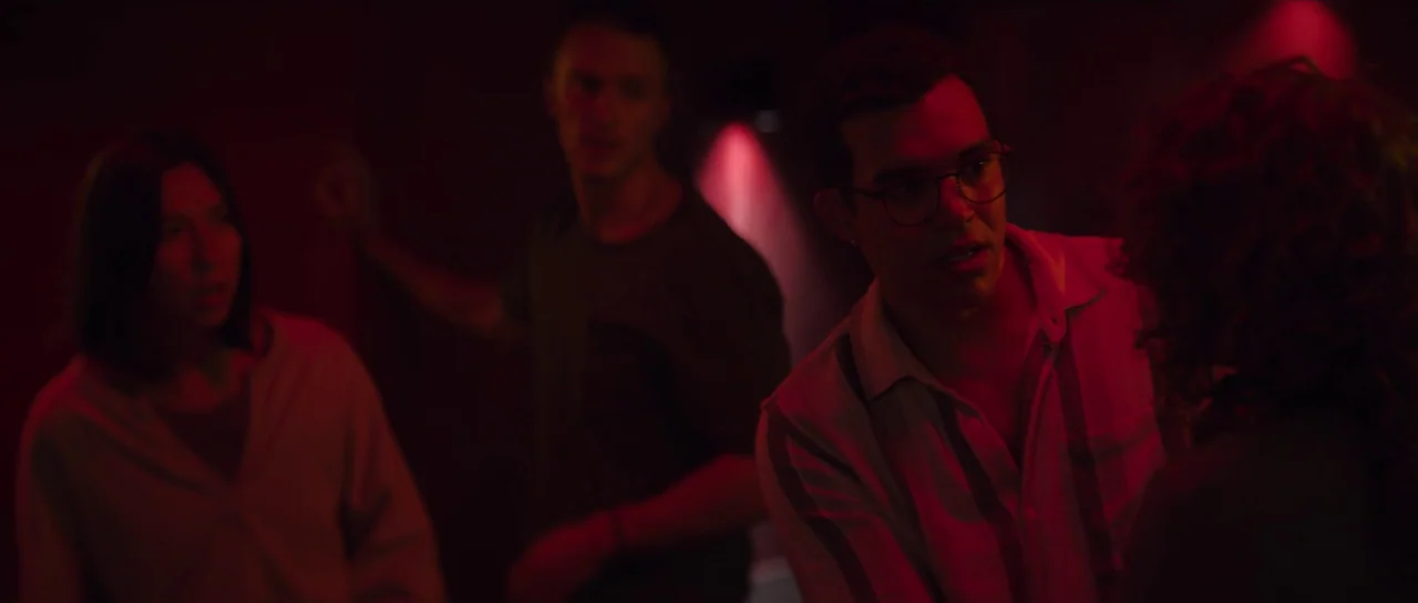 Hellraiser (2022) movie still: medium — A dark, red-lit nightclub environment with blurred silhouettes in the background. A group of three young…