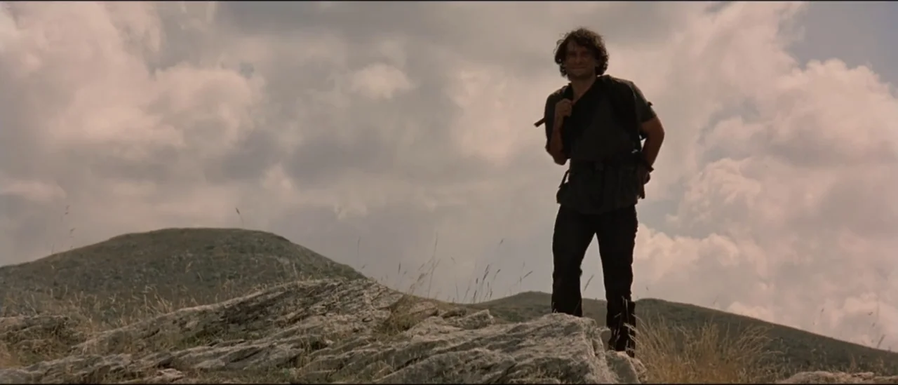 The Peacemaker (1997) movie still: fullbody — A man stands on a rocky mountain ridge under a sky filled with clouds. The subject is a man in casual…