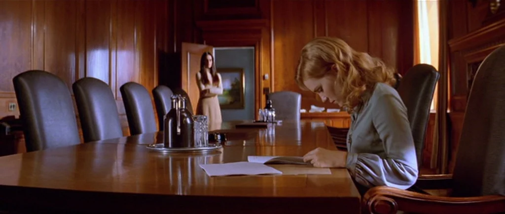 Where The Truth Lies (2005) movie still: medium — A paneled wooden conference room contains a long table with leather chairs. One woman sits reading…