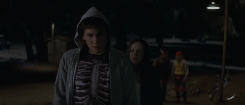 Donnie Darko movie still: medium — A dark residential street at night illuminated by a single overhead light. A young man in a skeleton shirt stands…