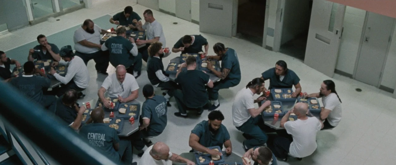Roofman (2025) movie still: wide — A utilitarian prison cafeteria features white floors, light walls, and several small metal tables. Numerous male…