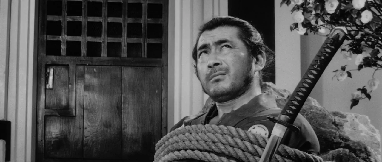 Sanjuro (1962) movie still: medium — A man stands in front of a wooden slatted door and a large stone. He is bound with thick ropes while looking…