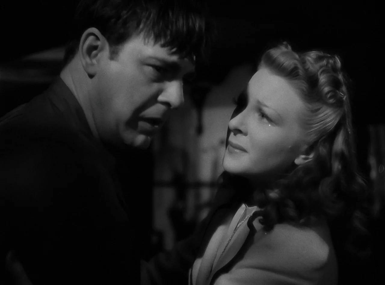 The Wolf Man (1941) movie still: medium — A dark indoor space with minimal architectural detail visible in the low-key lighting. An adult man and a…
