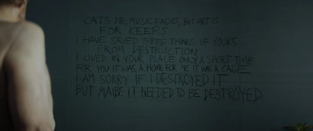 Inside (2023) (2023) movie still: medium — A dark, tiled wall features a block of handwritten text scrawled in black marker. A shirtless man stands…