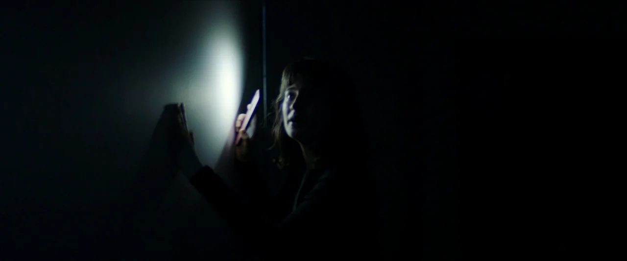 Double Blind (2023) movie still: medium — A dark, unlit room with minimal visual information other than a blank wall. A young woman holds an…