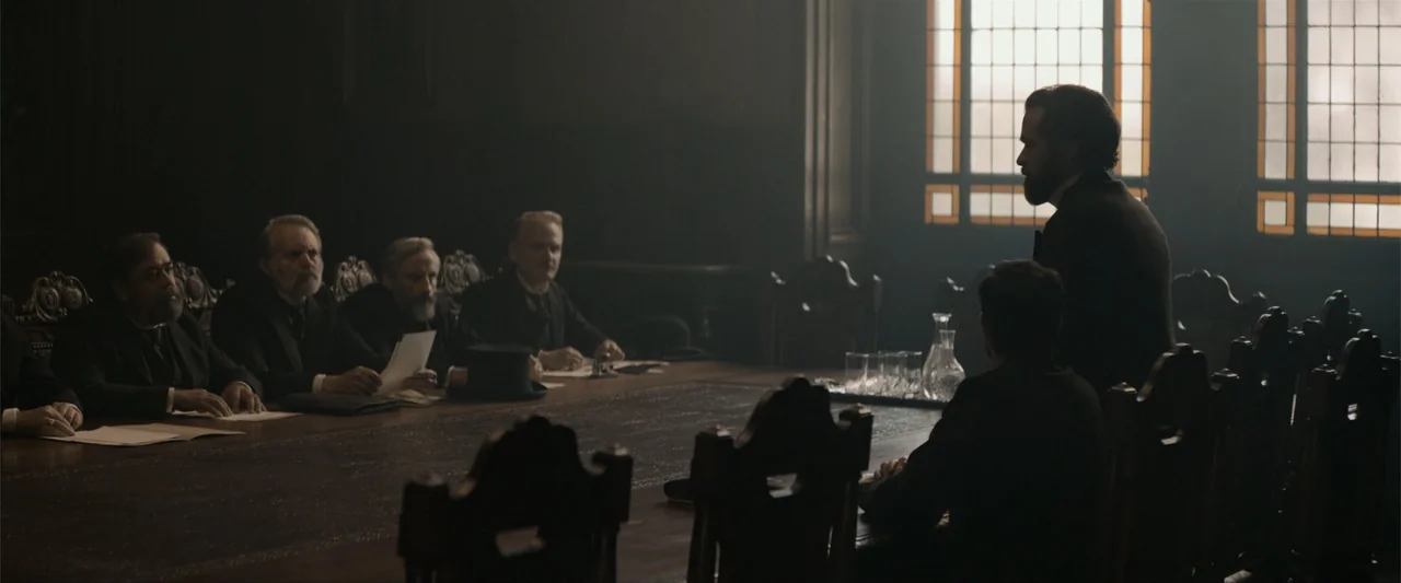 Eiffel (2021) movie still: wide — A dimly lit Victorian-era room contains a long heavy wood table with multiple chairs. A man stands at the table…
