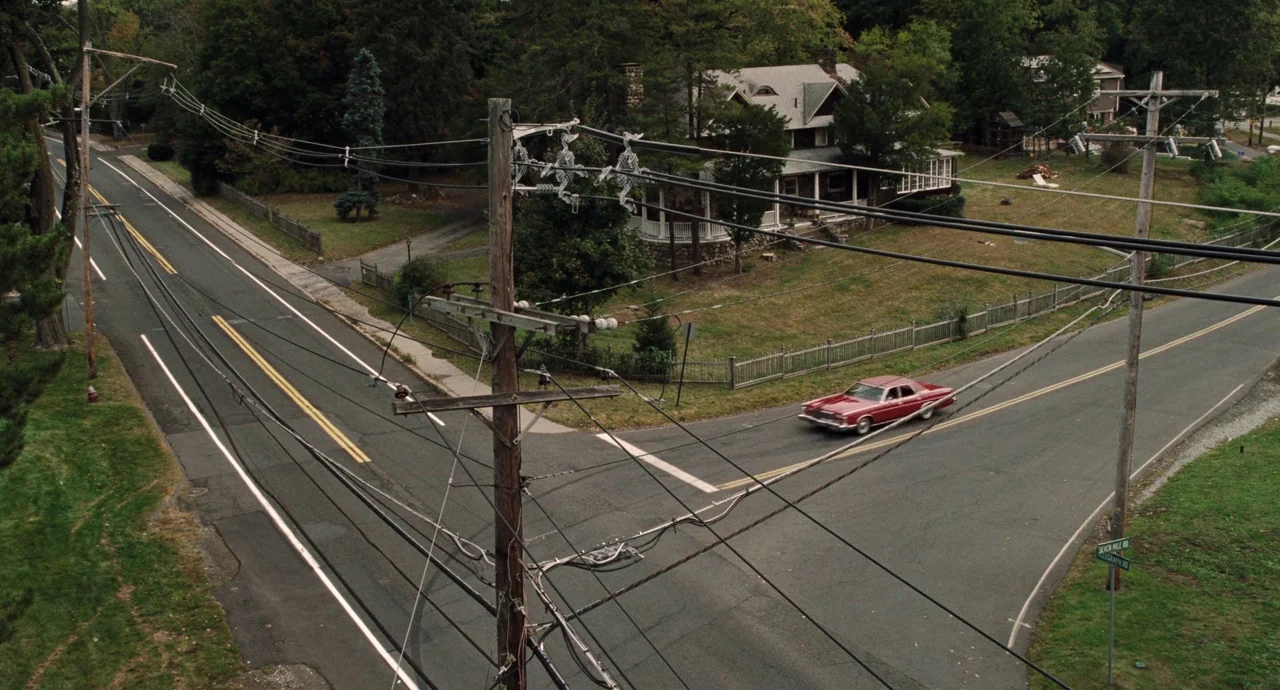The Irishman (2019) movie still: establishing — A quiet rural road intersection features power lines and a nearby house with trees. A vintage red…