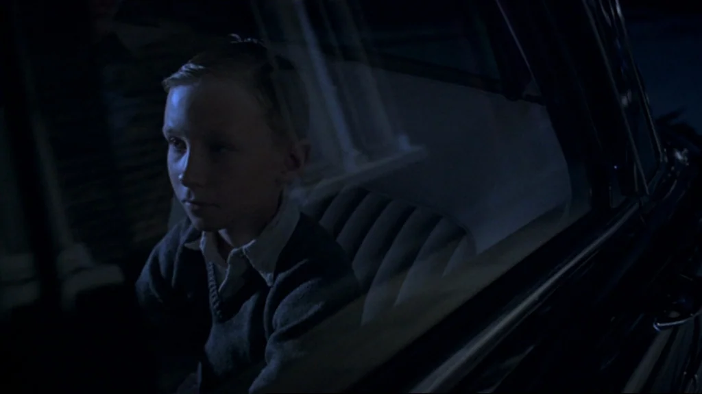 Spider (2002) movie still: medium — A young boy sits in the backseat of a vintage car at night. Blue light from the street enters through the window…
