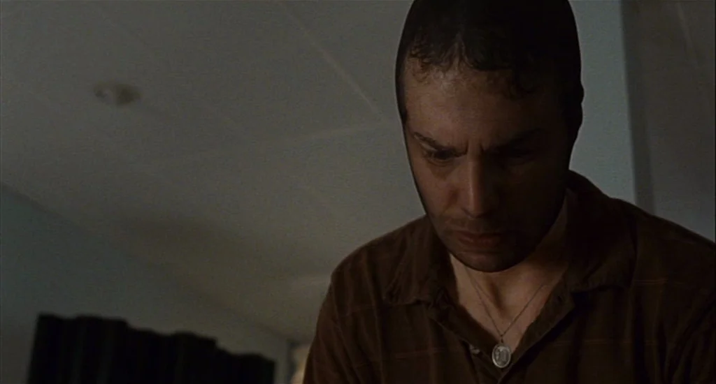 Choke (2008) movie still: closeup — A dimly lit indoor space with plain surfaces and shadows obscuring the surroundings. A man wearing a dark…