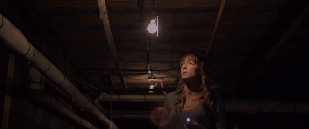 You’re Next (2013) movie still: medium — A dark subterranean space features exposed pipes and ceiling joists illuminated by a single bulb. A woman…