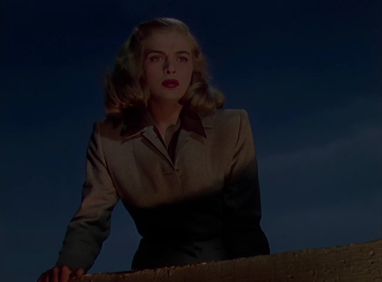 Desert Fury (1947) movie still: medium — A blonde woman in a brown blazer leans against a concrete ledge at night. The sky is dark and featureless…