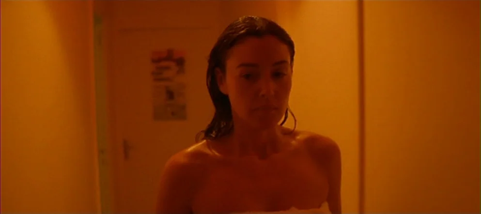 Irréversible (2002) movie still: medium — A narrow residential hallway is bathed in saturated orange artificial light. A woman walks forward wearing…