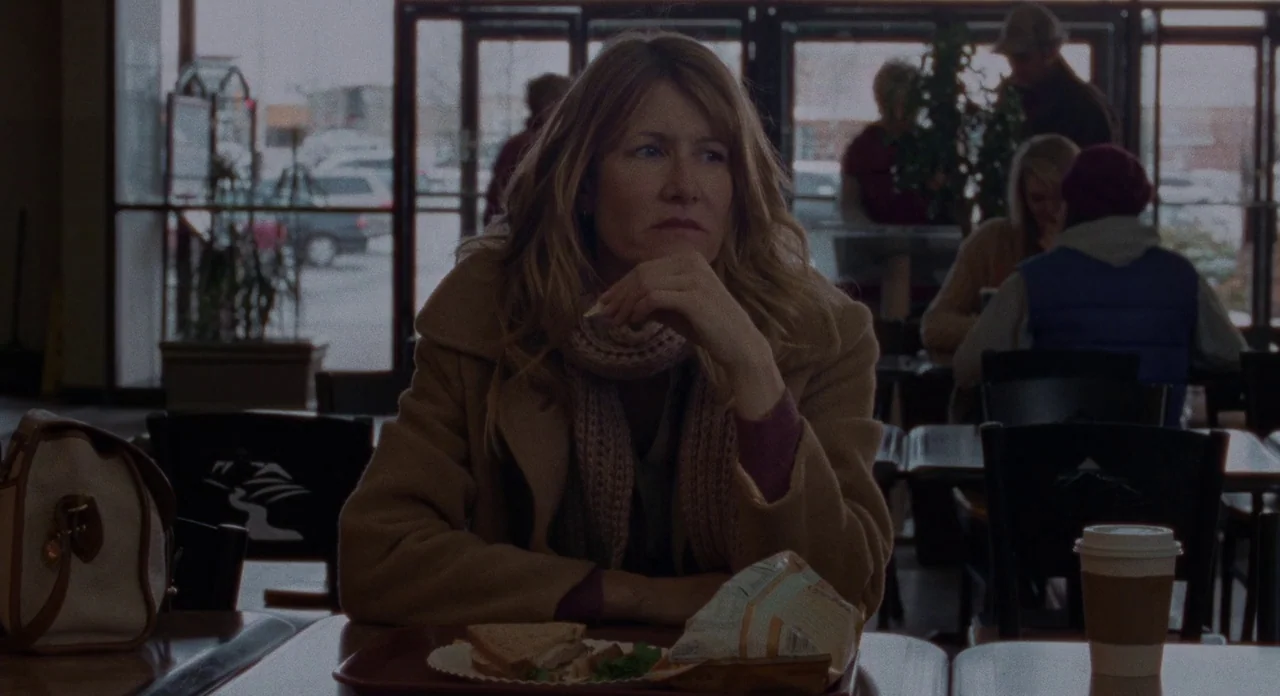 Certain Women (2016) movie still: medium — A quiet diner interior with large glass windows looking out onto a parking lot. A middle-aged woman sits…