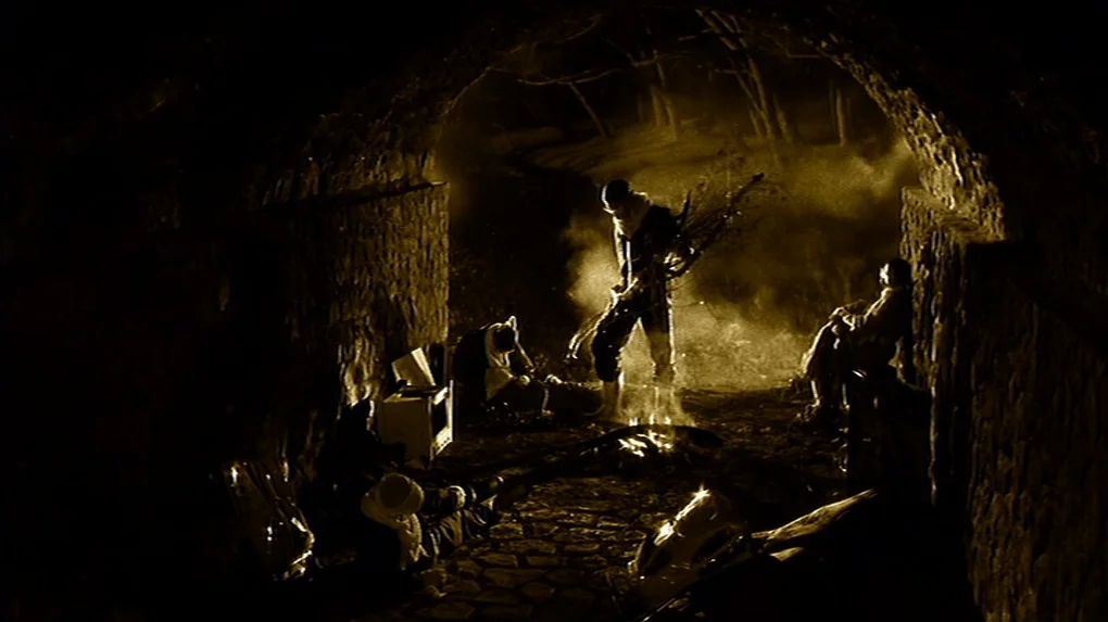 The State of Things (1982) movie still: wide — A group of people gathers around a campfire inside a dark stone tunnel at night. The fire casts light…