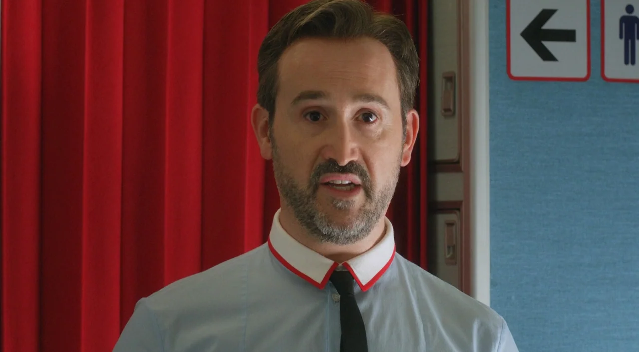 I’m So Excited! (2013) movie still: closeup — An airplane cabin interior features a red curtain and blue walls with signage. A man in a collared…