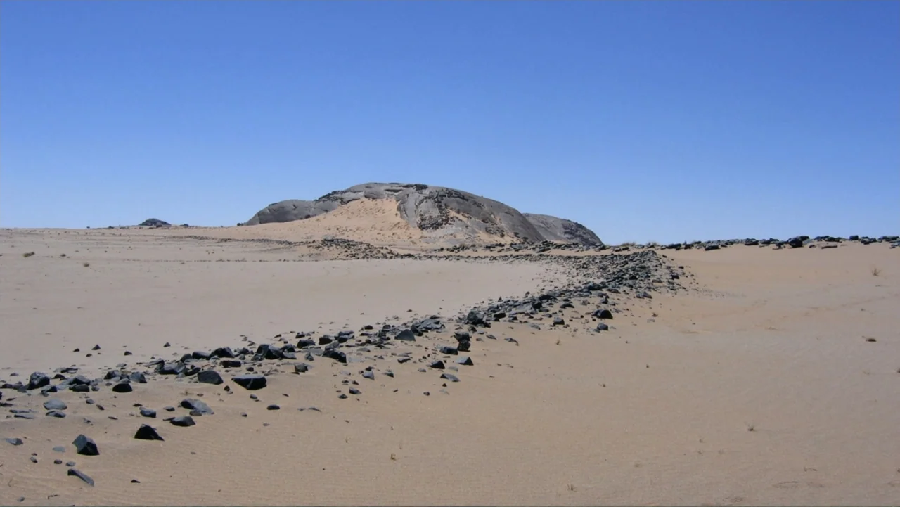 The Atlantis Puzzle (2024) movie still: establishing — An arid desert landscape features a vast expanse of sand scattered with dark rocks. A low,…