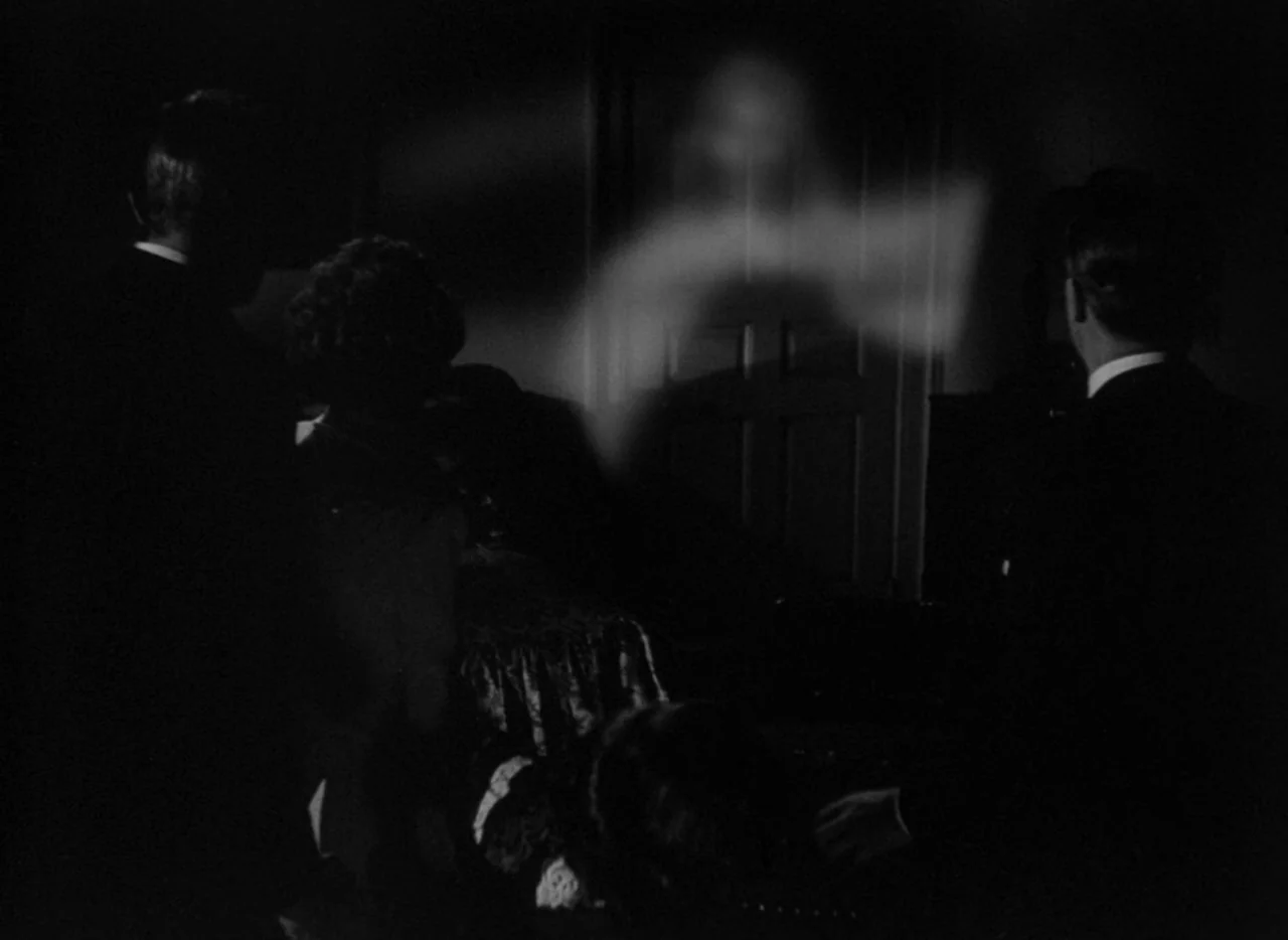 The Uninvited (1944) movie still: medium — A dark, dimly lit room contains three figures facing a partially transparent supernatural entity. The…
