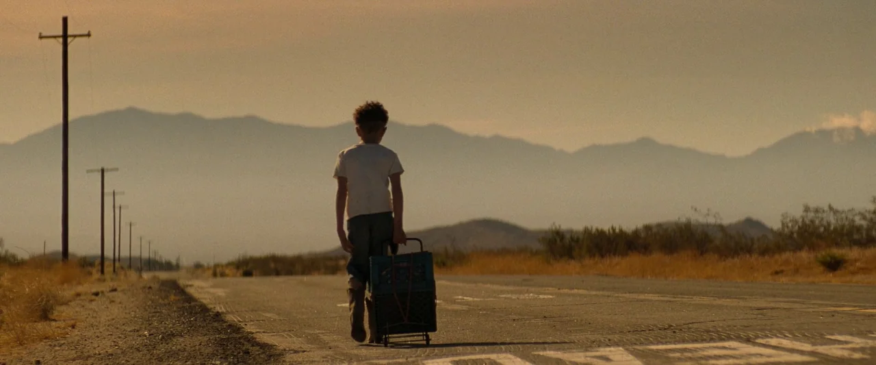 Bullet Train (2022) movie still: fullbody — A flat asphalt road stretches into a vast desert landscape with distant mountains. A young child walks…