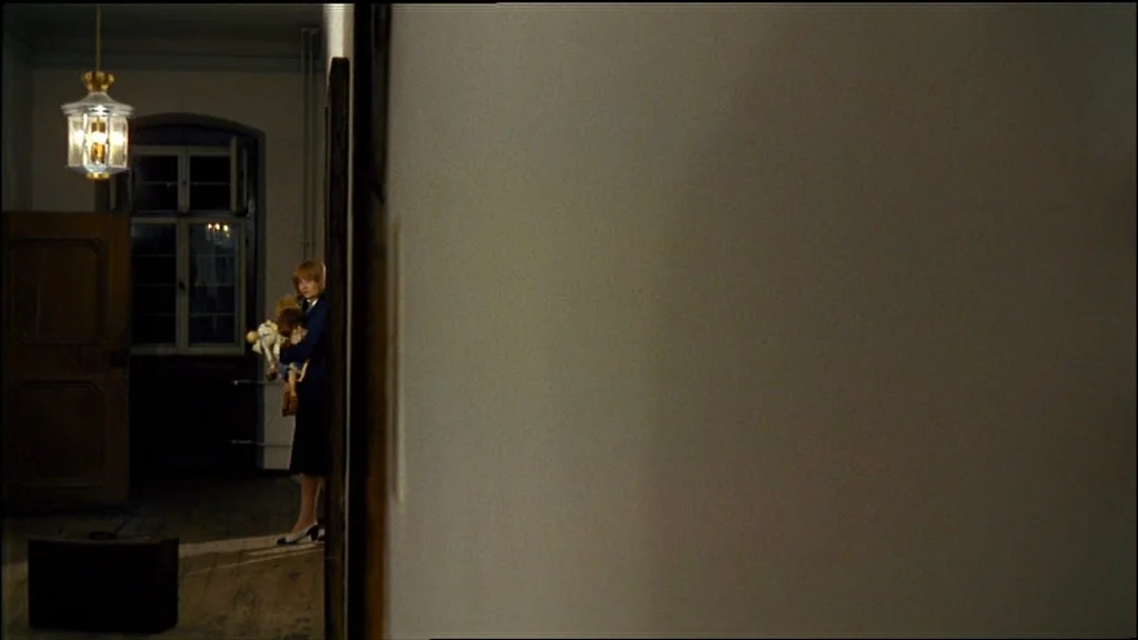 Chinese Roulette (1976) movie still: wide — A dimly lit, narrow corridor features a plain wall and an overhead lantern light. A young woman stands…