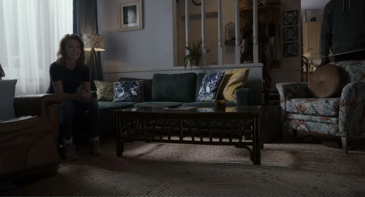 Gran Turismo (2023) movie still: medium — A residential living room contains a green corduroy sofa and a rattan coffee table. A woman smiles while…