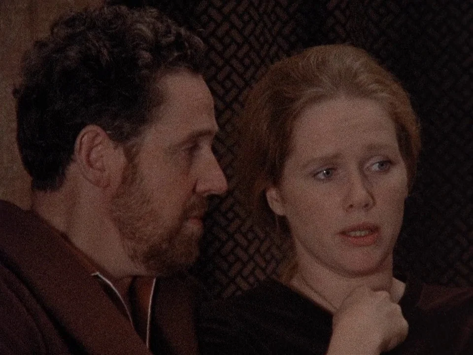 Scenes from a Marriage (1973) movie still: medium — A man and woman are positioned close to each other in a dimly lit space. The man watches the…