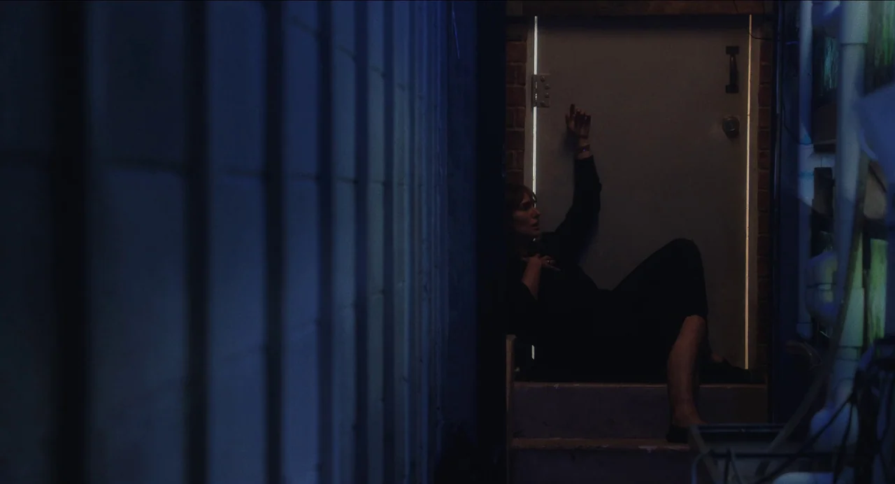 May December (2023) movie still: fullbody — A narrow dimly lit concrete stairwell with a door at the top. A woman sits on the bottom step reaching…