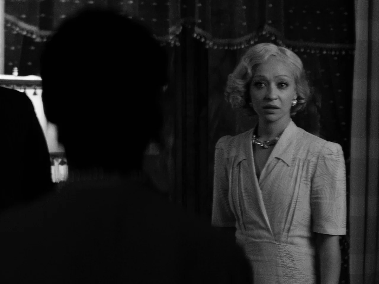 Passing (2021) movie still: medium — A dimly lit room with draped curtains provides a dark background for the subject. A blonde woman in a vintage…