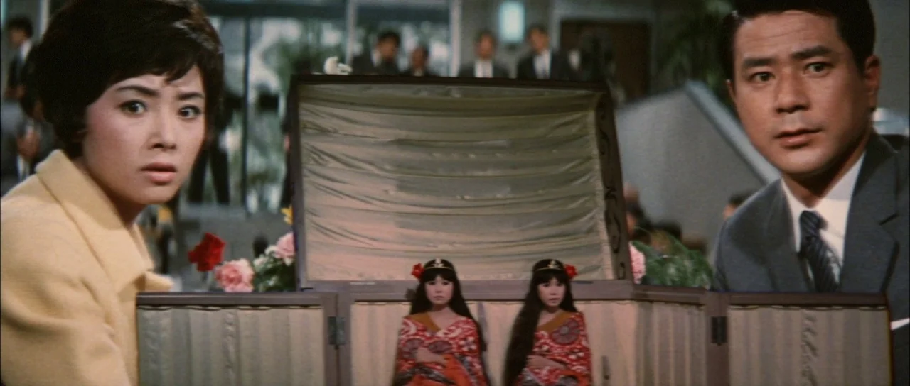 Ghidorah The Three Headed Monster (1964) movie still: medium — A well-lit lobby with blurry figures standing in the background. A woman and man look…
