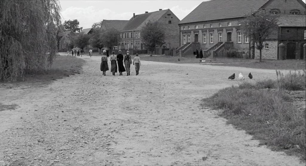 The White Ribbon (2009) movie still: wide — A rural village clearing features dirt paths between rustic brick buildings and trees. Five people walk…