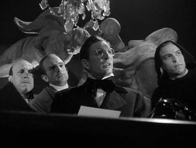 Mr. Arkadin (1955) movie still: medium — Four men sit at a table in a dimly lit room featuring a large stone sculpture. They look ahead with serious…