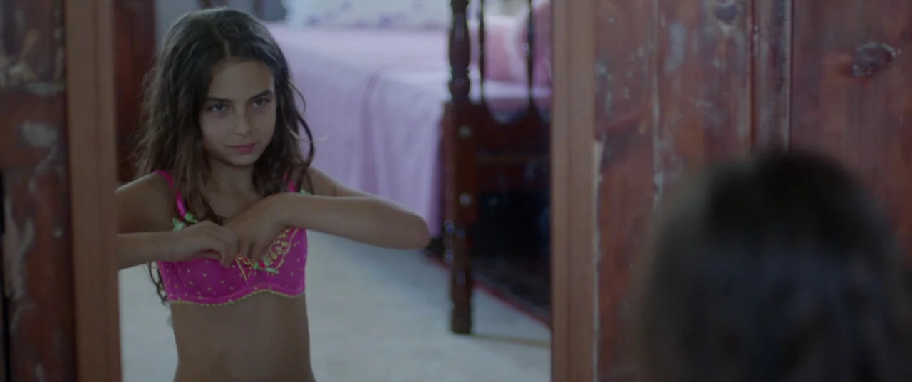 Mustang (2015) movie still: medium — A young girl stands in front of a full-length wooden mirror inside a room. She adjusts her pink bra while…