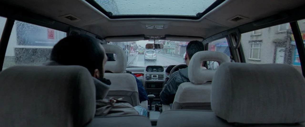 Catch Me Daddy (2014) movie still: medium — The interior of a moving car with two occupants viewed from the back seat. Rain droplets are visible on…